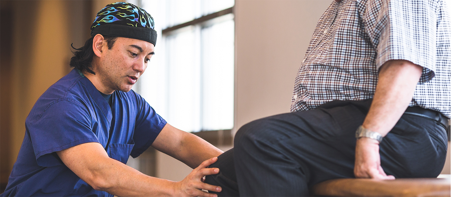 Male doctor wearing scrubs and a beanie cap examines male patient for knee replacement surgery.